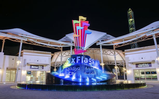 Six Flags Qiddiya City entrance with illuminated sign and colorful flags at night.