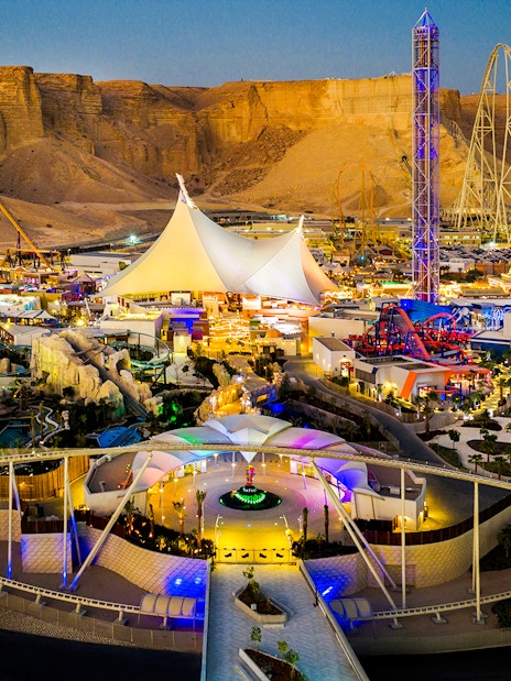 Aerial view of Six Flags Qiddiya City with illuminated rides and attractions against a desert backdrop.